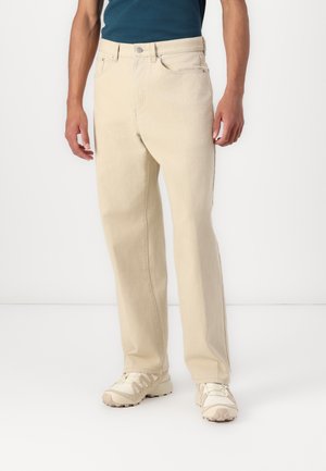 Ljust beige jeans med rak passform, femfickorsdesign och subtila sömdetaljer. Bärs med en blå skjorta och beiga sneakers.