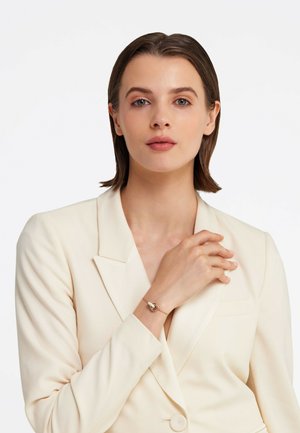 Bracelet en or et en argent avec un pendentif circulaire, porté au poignet. Le modèle porte un blazer crème en tissu texturé. Design simple et élégant.