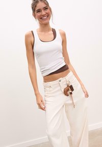 Femme souriante en débardeur blanc et tee-shirt marron, portant un pantalon blanc ample avec un porte-clés accroché, sur un fond clair.