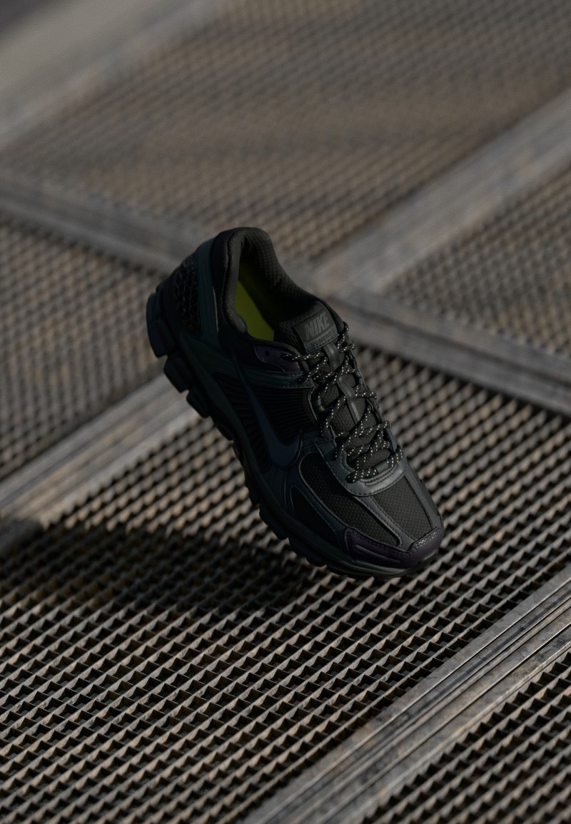 Chaussure de course Nike noire avec des lacets réfléchissants posée sur une surface métallique à grille avec un motif de grille diagonale.