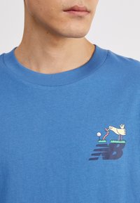 Zila kokvilnas t-kreklu ar ribotu apaļu kakla izgriezumu, kurā ir attēlots karikatūras putns un tārps virs tumši zilā New Balance logo.