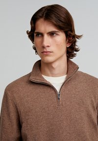 Brun zip-up sweater lavet af tekstureret strikstof, med høj krave, lynlåslukning og diskret brandingdetalje på brystet.