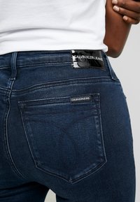 Tmavě modré džíny s přiléhavým střihem, mají černou nášivku s nápisem "Calvin Klein Jeans" a výrazné švy na kapsách.