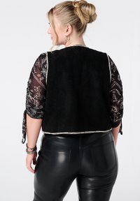 Zwart suède vest met een zilveren rand, gecombineerd met een doorschijnende zwarte mouw met bloemenpatroon. Draagt een glanzende zwarte leren broek.