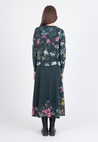 Floral-knit long dress in dark green, featuring multi-colored flower patterns at the hem, long sleeves, and a relaxed fit. Worn with black boots.