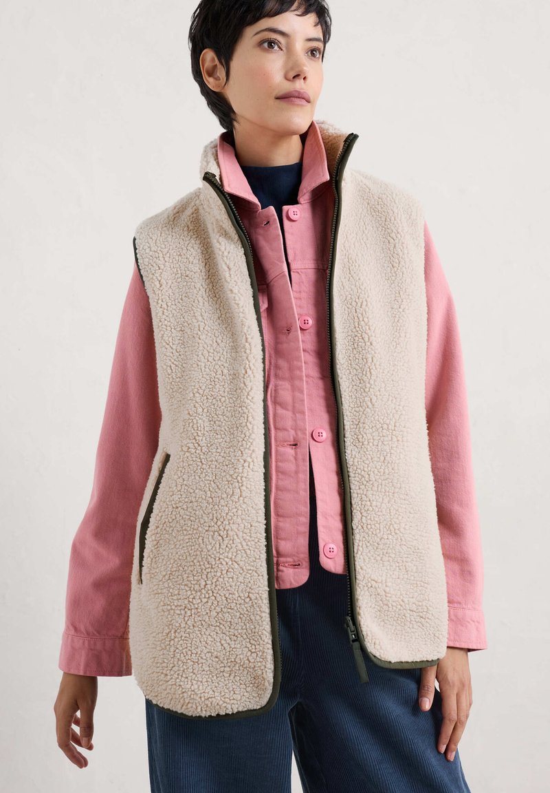 Fleece vest in crèmekleur met groene accenten, voorzien van zijzakken en een hoge kraag, gedragen over een roze overhemd met knopen. Gelaagd, warm materiaal.