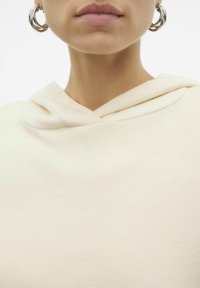 Felpa color crema con una texture morbida e un dettaglio del cappuccio ripiegato. Accentuata da grandi orecchini circolari d'argento. Inquadratura ravvicinata.