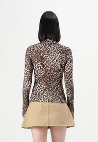 Persoon met steil zwart haar, gekleed in een langärmige top met luipaardprint en een tan minirok, staande met de rug naar de camera op een witte achtergrond.