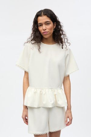 Žena s kučeravými tmavými vlasmi nosiaci krémový krátky rukáv s volánikovým lemom a zodpovedajúce šortky na svetlom jednofarebnom pozadí.