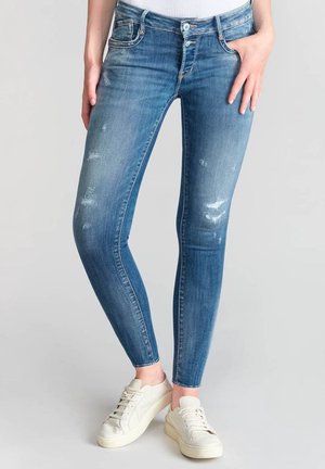 Kobieta nosząca niebieskie obcisłe, postrzępione dżinsy i białe trampki, stojąca na tle jednolitego jasnoszarego tła.