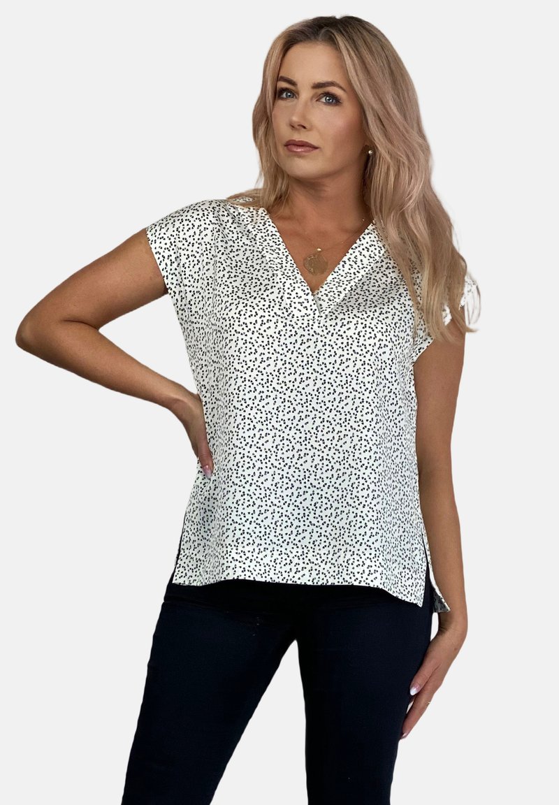 Biała bluzka z dekoltem w kształcie litery V, krótkimi rękawami i czarnym wzorem w kropki. Lekki materiał z luźnym krojem. Noszona z ciemnymi spodniami.