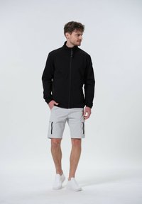 Schwarze Zip-Jacke mit hohem Kragen, kombiniert mit hellgrauen Cargo-Shorts mit Reißverschlusstaschen. Das Model trägt weiße Turnschuhe.