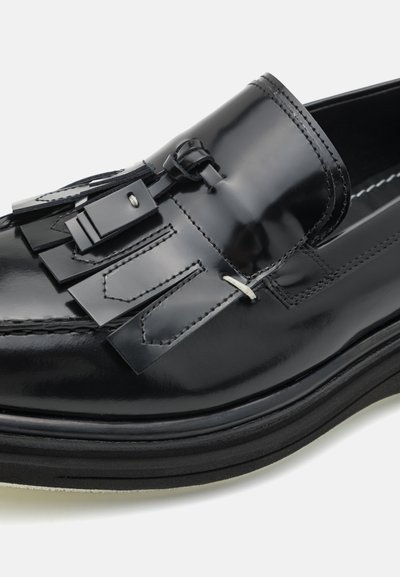 Mustat kiiltonahasta valmistetut loafer-kengät, joissa on koristeelliset fringet ja metalliosat. Pyöreä kärki ja erottuva musta pohja. Suloinen pinta.