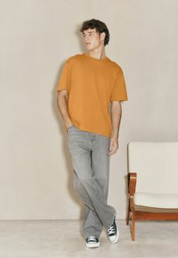 Oranges Kurzarm-T-Shirt mit lockerer Passform, kombiniert mit hellgrauen Jeans und schwarzen Sneakers, steht neben einem Stuhl aus hellem Holz.
