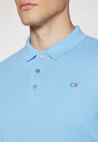 Svijetloplavi polo majica od teksturiranog materijala, s klasičnom ovratnikom, tri siva gumba i malim vezom CK logotipa na prsima.