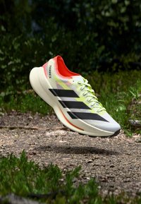 Weißer Sportschuh mit schwarzen Streifen, einem roten Innenfutter und neon-grünen Schnürsenkeln. Leichtes Design und strukturierte Oberseite für besseren Halt.