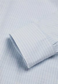Dettaglio di una camicia azzurro chiaro con sottili righe bianche. Presenta un polsino ben rifinito con cuciture a vista, che mette in mostra una texture di cotone morbida.