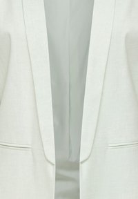 Bela blazer z ovratnikom šal in dvema horizontalnima režastima žepoma, poudarek na sprednjem odprtju in reverjih.