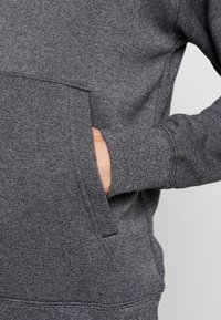 Grå sweatshirt med texturerad tyg, känguruficka och ribbade ärmar. Närbild av en hand som vilar i fickan.