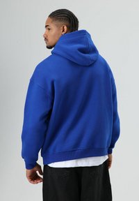 Blauwe katoenen hoodie met een ontspannen pasvorm, een grote capuchon en geribbelde manchetten. De achterkant toont een gladde textuur en strakke lijnen.