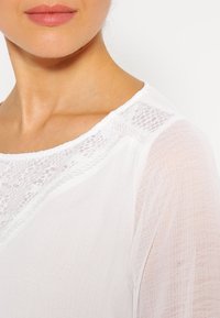 Blusa bianca con un bordo in pizzo lungo lo scollo. Il tessuto è trasparente e strutturato, con una finitura liscia. Inquadratura ravvicinata dello scollo.