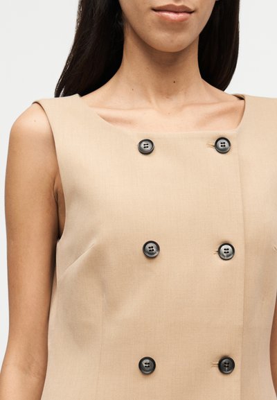 Beiges ärmelloses Kleid mit quadratischem Ausschnitt, ausgestattet mit vier schwarzen Knöpfen auf der Vorderseite und einer glatten Stofftextur.