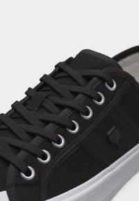 Μαύρο πάνινο αθλητικό παπούτσι με μαύρα κορδόνια περασμένα από ασημένιες τρύπες και λευκή λαστιχένια σόλα, με ένα μικρό μαύρο έμβλημα στο πλάι.