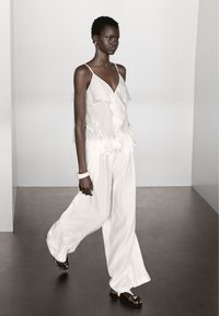 Witte, gerimpelde mouwloze top met delicate stofdrapering, gecombineerd met brede witte broek en zwarte sandalen, gedragen door een model dat loopt.