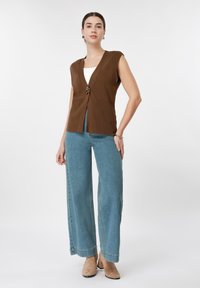 Γυναίκα που φοράει χωρίς μανίκια καφέ γιλέκο πάνω από λευκό τοπ, ανοιχτό μπλε παντελόνι φαρδιάς γραμμής, μπεζ μποτάκια αστραγάλου και χρυσούς κρίκους σκουλαρίκια.