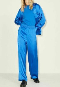 Satin blue tracksuit with a fitted top and loose pants. Features an elastic waistband and long sleeves, paired with black shoes.