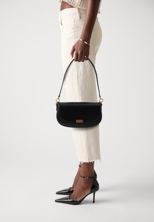 Sac en cuir noir au design arrondi, avec des accessoires en or et une courte poignée, tenu par une personne portant un pantalon clair et des escarpins noirs.