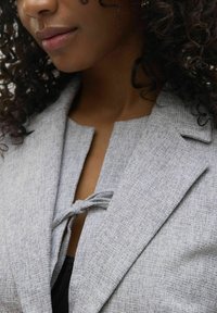 Blazer gris claro con tejido texturizado, que presenta una abertura en forma de ojo de cerradura en el cuello y un detalle atado. Solapas con muesca en el cuello.