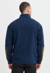 Veste en polaire bleu marine avec des accents vert olive sur les manches, col haut et coupe décontractée. Finition texturée, sans fermetures visibles.