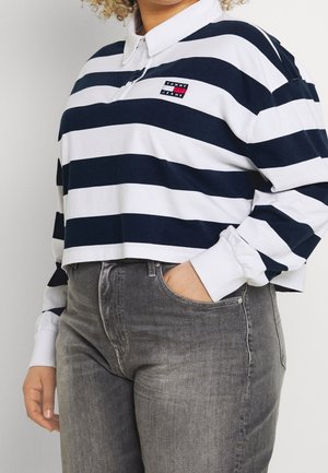 Persoon draagt een marineblauw-wit gestreept shirt met lange mouwen, een witte kraag en grijze spijkerbroek, hand in de zak.