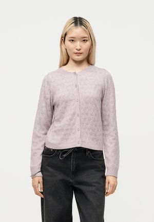 Ung kvinde iført en lyslilla knappet cardigan med et subtilt mønster og sorte højtaljede jeans, stående mod en ensfarvet baggrund.