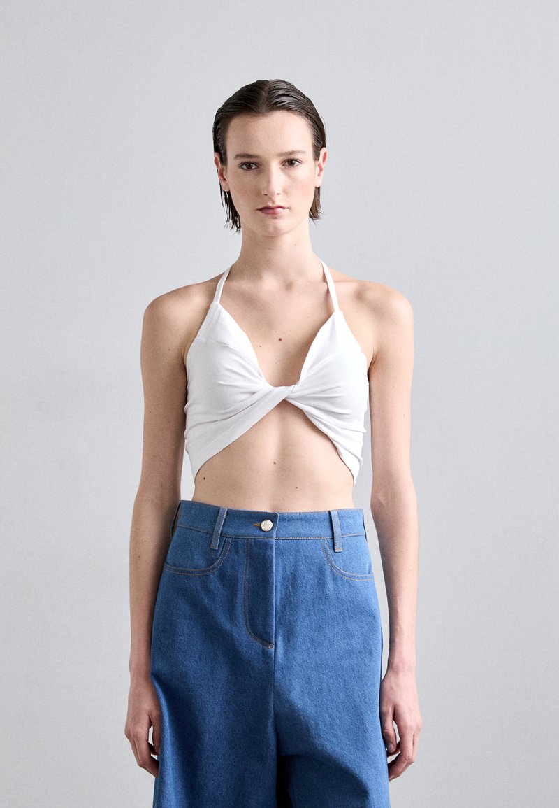 Weißes Trägeroberteil mit verdrehtem Vorderdesign, kombiniert mit hochgeschnittenen blauen Jeanshosen mit Fronttaschen und orangen Nähten.