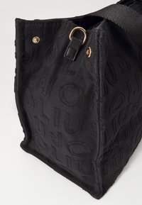 Sac cabas en tissu noir avec motif de texte en relief, anneau en métal doré et fermeture à bouton-pression, et anses épaisses noires.