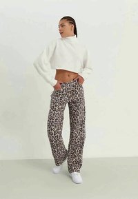 Sweater branco cropped com gola alta e mangas longas; calças beges com padrão de leopardo preto; ténis brancos; modelo a posar contra um fundo claro.