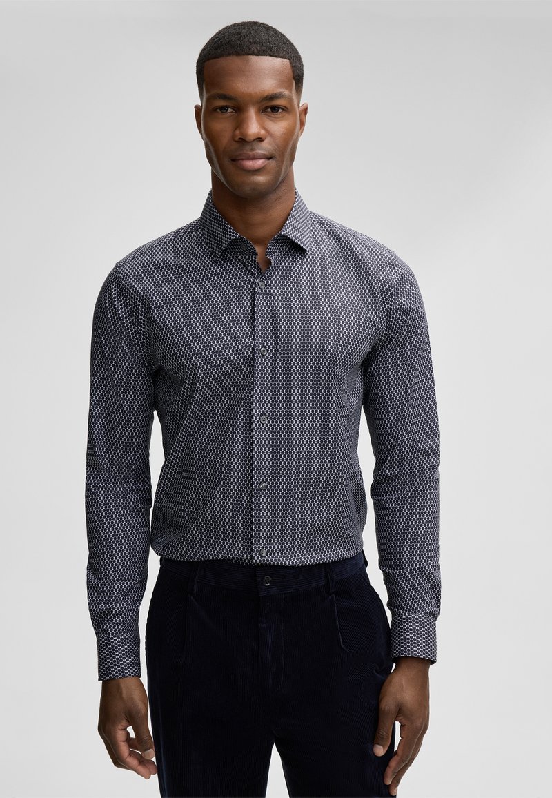 Camicia blu con bottoni, caratterizzata da un motivo geometrico bianco, maniche lunghe e colletto standard, abbinata a pantaloni di velluto a coste scuri.