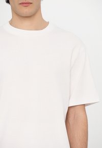 Jeune personne portant un t-shirt blanc uni à manches courtes avec col rond, visible du cou jusqu’au milieu du torse sur fond blanc.