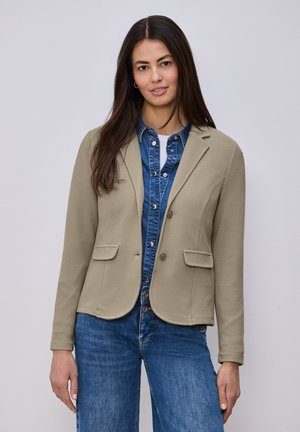Femme aux longs cheveux noirs portant un blazer beige sur une chemise en denim bleue boutonnée et un jean bleu, debout devant un fond gris uni.