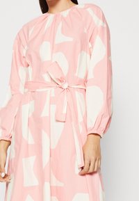 Růžové šaty vyrobené z lehké tkaniny, s bílými abstraktními tvary, dlouhými rukávy s elastickými manžetami a opaskem na zavazování v pase.