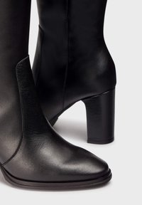 Botines de tacón alto de cuero negro con punta afilada, textura suave y paneles elásticos laterales. Presentan un sólido tacón cuadrado.