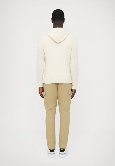 Voľná krémová mikina s kapucňou s textúrovaným pleteným vzorom, doplnená béžovými cargovými nohavicami s bočnými vreckami a bielymi teniskami.