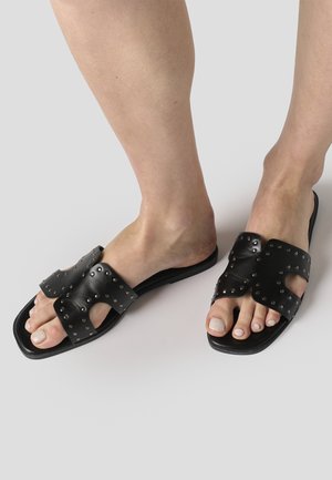 Pies con sandalias tipo slide de cuero negro con tachuelas de metal y diseño recortado, sobre un fondo liso de color gris claro.