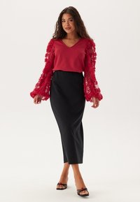 Rød top med bølgende, gennemsigtige ærmer, parret med en sort tætsiddende nederdel og sorte sandaler med blomsterdetaljer. Glat stofstruktur.