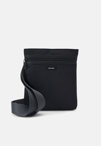 Schwarze Stoff-Schultertasche mit einem Reißverschluss, glatter Textur und einem grauen verstellbaren Tragegurt. Auf der Vorderseite befindet sich ein kleines Logo-Patch.