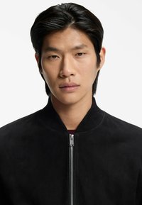 Black sueded bomber jacket with a ribbed collar and full zipper. The fabric has a soft texture, offering a clean and minimalistic design.