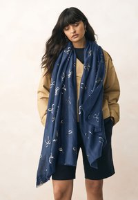 Sciarpa blu navy in tessuto strutturato, con un motivo ricamato in oro di anelli e fiocchi. Drappeggiata casualmente su una giacca color cammello.