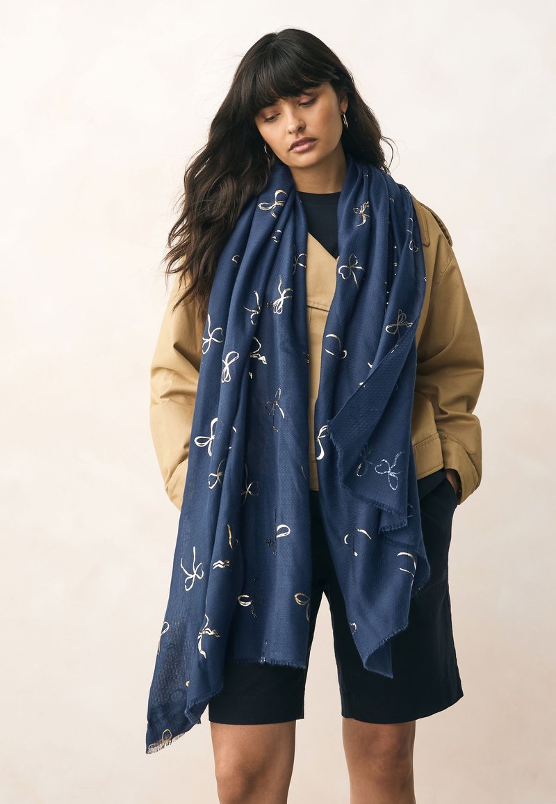 Sciarpa blu navy in tessuto strutturato, con un motivo ricamato in oro di anelli e fiocchi. Drappeggiata casualmente su una giacca color cammello.